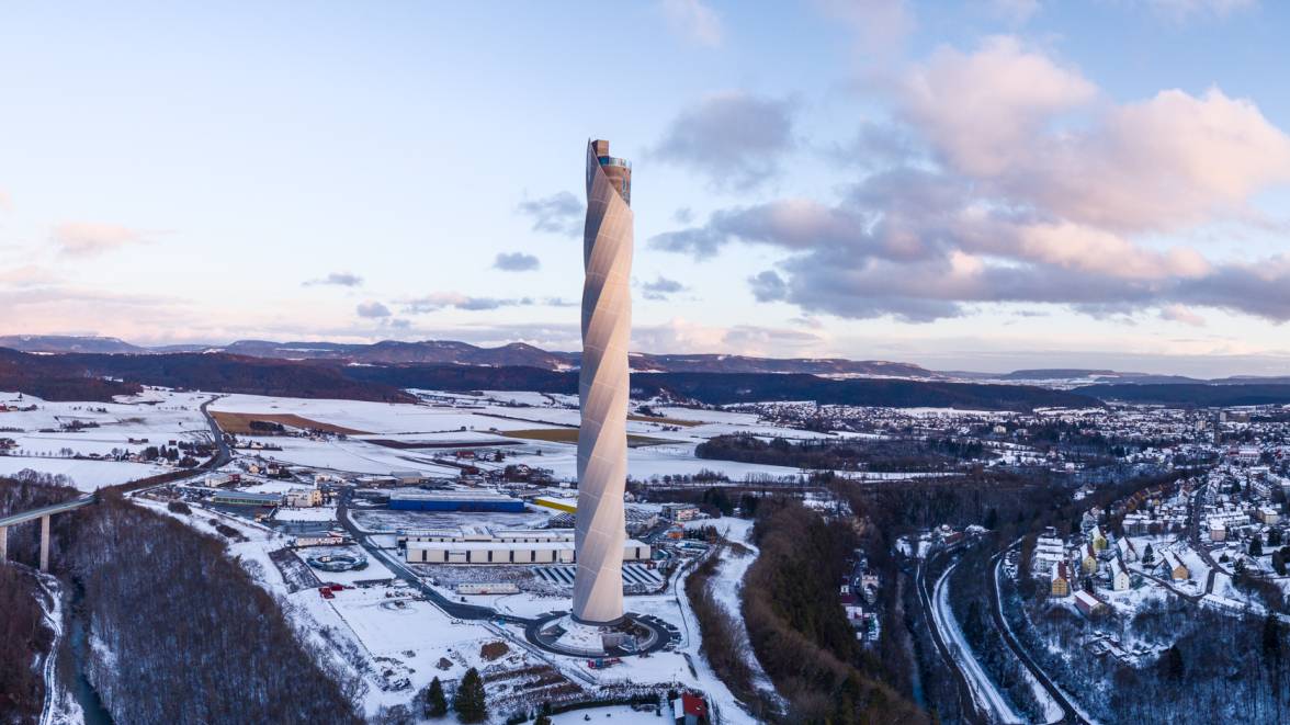 Eine winterliche Landschaft mit schneebedeckten Bäumen und einem hohen, schlanken Testturm für Aufzüge, der in den Himmel ragt. 