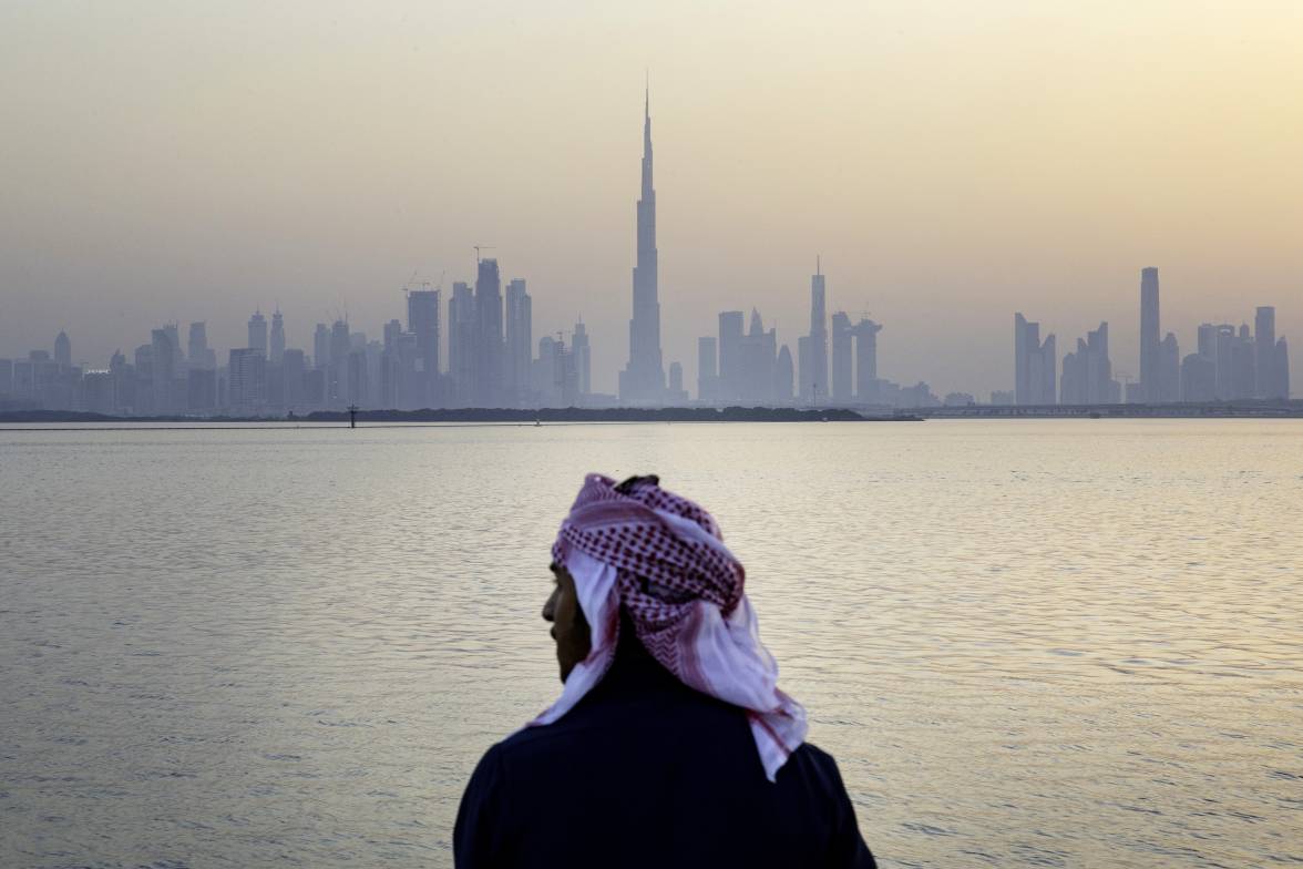 Eine männliche Figur in traditioneller arabischer Kleidung, die von hinten vor einer Wasserfläche abgebildet ist. Im Hintergrund ist die Skyline von Dubai zu erkennen. 